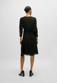 Schwarzes Midikleid aus strukturiertem Stoff, mit Puffärmeln, Kordelzug in der Taille und gestuftem Rock. Abgeschlossen mit einem Schlüsselrücken und geknöpften Manschetten.