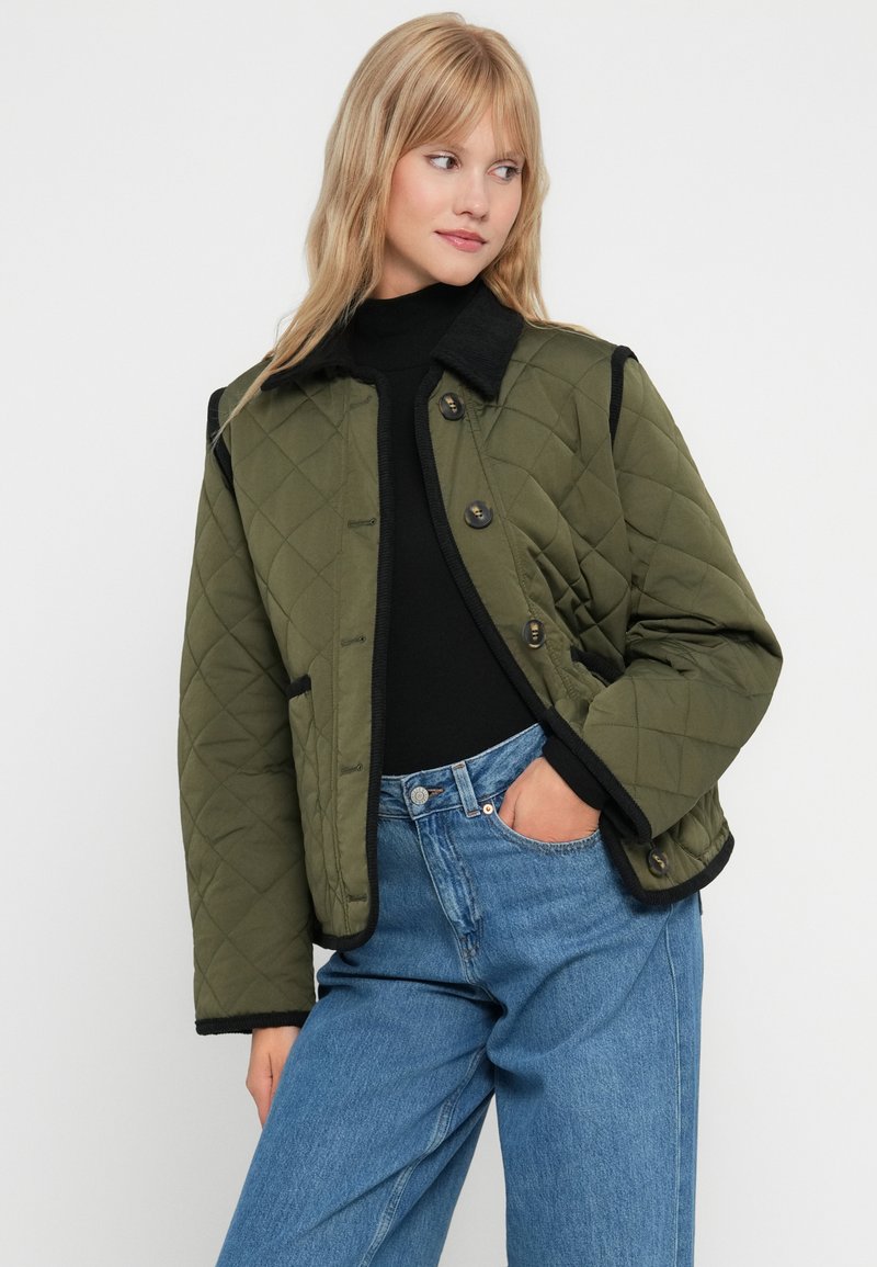 Quilted olivgrüne Jacke mit schwarzen Akzenten, mit Knopfverschluss, Kragen und Seitentaschen, getragen über einem schwarzen Top und blauen Jeans.