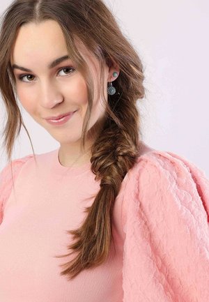 Vrouw met lang, bruin haar in een vlecht, gekleed in een lichtroze top met zachte mouwen en ronde oorbellen. Schone achtergrond.