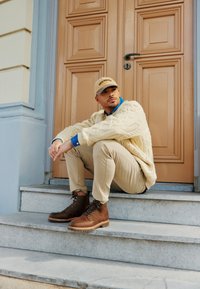 Maglione a maglia beige, camicia a colletto blu sotto, pantaloni beige, stivali in pelle marrone con suole in gomma, seduto su gradini di pietra grigia accanto a una porta color beige.