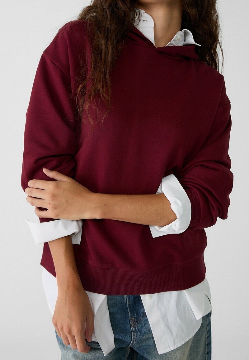 Personne portant un sweat-shirt bordeaux par-dessus une chemise blanche à manches retroussées et un jean bleu, les bras croisés sur le torse.