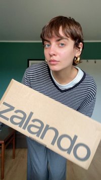 Karton doos met het woord "zalando" in grote grijze letters. Een persoon in een gestreepte top houdt de doos vast in een goed verlichte binnenruimte.