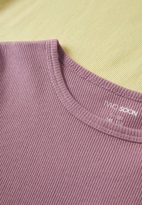 Μωβ ριμπ πλεκτό μπλουζάκι με στρογγυλή λαιμόκοψη, απλωμένο σε μπεζ επιφάνεια, που δείχνει τις λεπτομερείς ραφές και την ετικέτα με το λογότυπο μέσα στο γιακά.