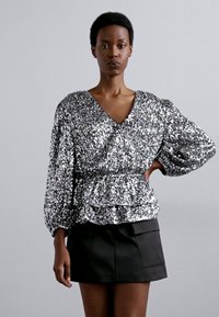 Sudraba sequinu tops ar V formu izgriezumu, puffētiem garām piedurknēm un savilktā pepluma jostu, komplektēts ar melnu miniskirti.