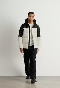 Puffjakke med et tofarvet design bestående af en hvid krop og sorte skuldre, parret med sorte bukser og en hvid ribstrikket sweater.