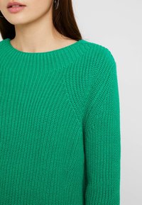 Zielony sweter z dzianiny o teksturowanej, prążkowanej tkaninie, z szerokim okrągłym dekoltem i minimalistycznym designem, przedstawiony od ramienia w górę.
