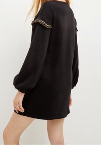 Svart sweatshirtklänning med långa puffärmar och rynkning med guldaccenter längs axlarna. Mjukt, texturerat tyg och avslappnad passform.