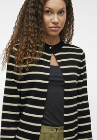 Melnā un krēmveida svītraina kardigāna ar apaļu apkakli, priekšējo pogu aizdari un atvērtas kakla izgriezuma dizainu, izgatavots no mīksta audumu maisījuma.