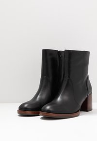 Botines de cuero negro con un ligero brillo, con cremallera lateral, punta redondeada y tacón bloque de madera. Diseño liso y elegante.