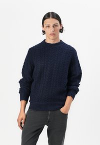 Suéter de punto trenzado en azul marino, hecho de hilo texturizado, con un escote redondo y puños y dobladillo acanalados. Combinado con jeans oscuros.