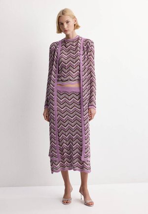 Femme portant une jupe en maille chevron violet, marron et blanc, un haut à manches longues assorti et un long cardigan ouvert devant avec des sandales à talons hauts.