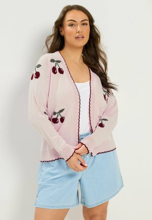Kvinde iført en lyserød cardigan med kirsebærmønster og bordeaux kant, hvid top og lyseblå denimshorts, stående foran en ensfarvet baggrund.
