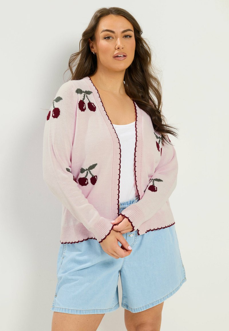 Kvinde iført en lyserød cardigan med kirsebærmønster og bordeaux kant, hvid top og lyseblå denimshorts, stående foran en ensfarvet baggrund.
