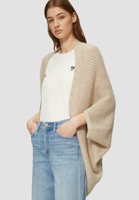 Beige gestrickte Cardigan mit offenem Vorderteil und weiten Ärmeln, getragen über einem weißen T-Shirt und hellblauen Jeans, mit lockerer Passform und strukturiertem Design.