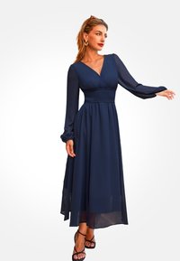 Granatowa sukienka o średniej długości, z marszczonym pasem, dekoltem w serek i długimi bufiastymi rękawami, wykonana z lekkiego, zwiewnego materiału. Czarne sandały na paski.