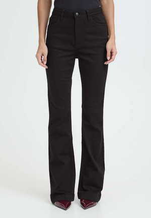 Kvinde iført højtaljede sorte flared jeans og burgunder spidse sko, stående mod en ensartet lys baggrund.