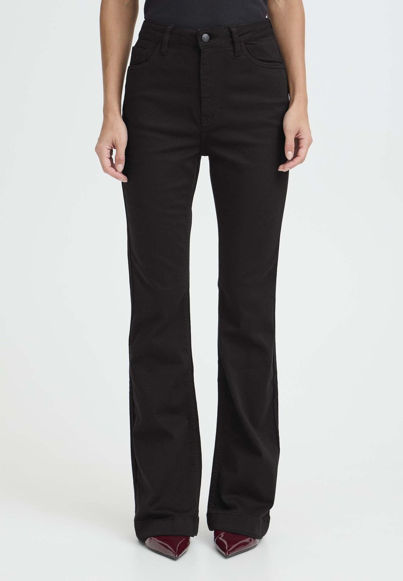 Mujer vestida con pantalones acampanados negros de cintura alta y zapatos burgundy de puntera afilada, de pie contra un fondo claro y sencillo.