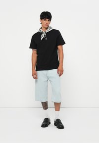 Camiseta de algodón negra con un logo en el frente, pantalones cortos de mezclilla azul claro, zapatos negros sin cordones y una bufanda de cuello blanca con patrón negro.
