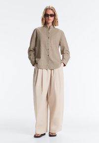 Femme portant une chemise à carreaux marron boutonnée, un pantalon beige à jambes larges, des sandales noires et des lunettes de soleil foncées, debout les mains dans les poches.