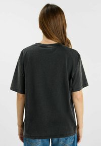 Mujer con cabello largo castaño viste una camiseta negra de gran tamaño y descolorida y jeans azules, de pie de espaldas sobre un fondo blanco liso.