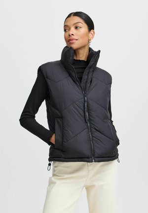 Black puffer vest with high collar, diamond quilted design, zip front, and side pockets. Worn over a long-sleeve black top and cream pants.