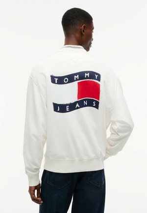 Mann trägt weißen Sweatshirt mit großem rot-weiß-blauem Tommy Jeans Logo auf dem Rücken, kombiniert mit dunkelblauer Jeans.