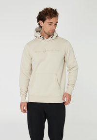 Sudadera beige hecha de tejido suave, con un bolsillo frontal, capucha con cordones y logo grabado de "SAIL RACING" en el pecho.