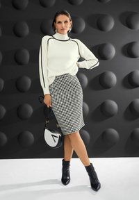 Frau, die in einem weißen Pullover mit schwarzen Streifen, einem Schwarz-Weiß-Hahnentrittmuster-Rock, schwarzen Stiefeletten posiert und eine schwarz-weiße Handtasche hält.