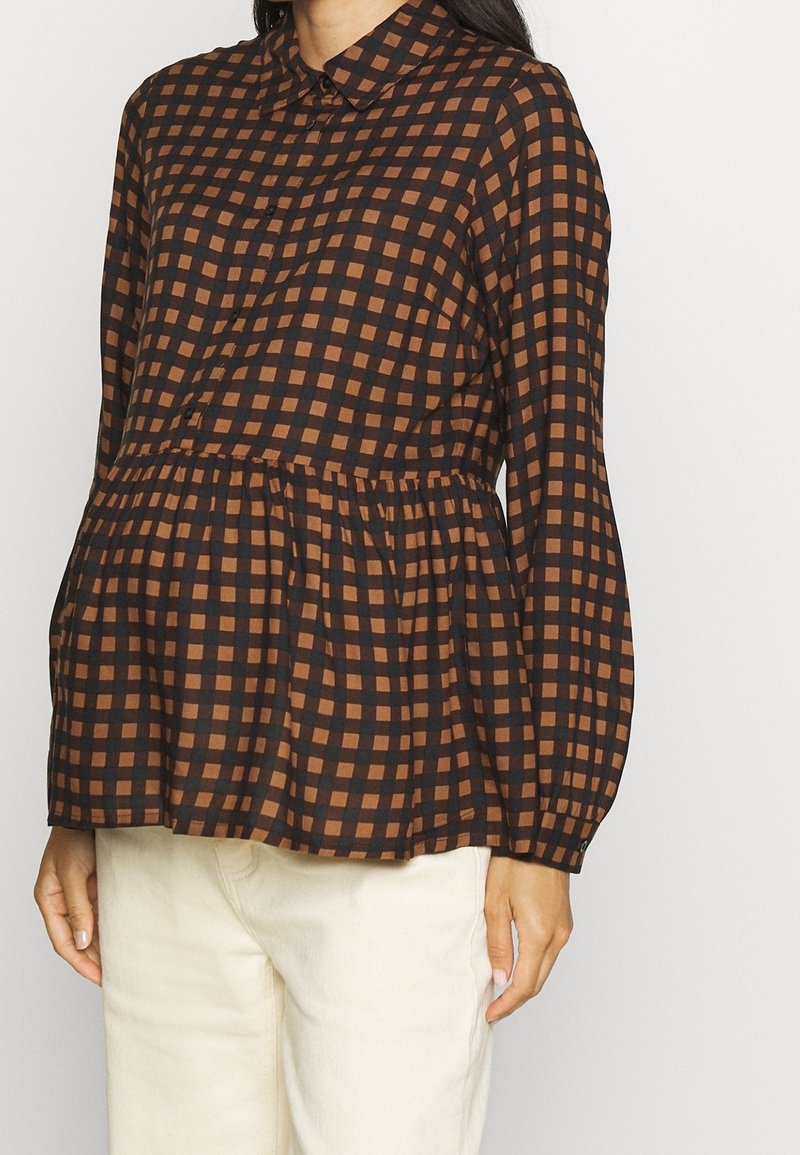 Blouse à carreaux marron et noir avec col boutonné, taille froncée et manches longues, présentant une coupe décontractée et un tissu léger.