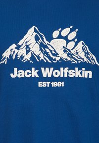 T-shirt en coton bleu avec un graphisme blanc représentant des montagnes, une empreinte de patte, et le texte "Jack Wolfskin EST 1981" au centre.