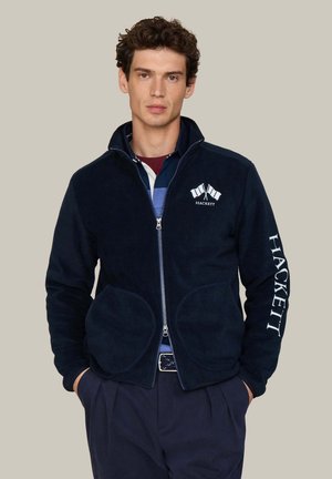Junger Mann trägt eine marineblaue Jacke mit Reißverschluss und Hackett-Logo auf Brust und Ärmel, steht mit den Händen in den Taschen vor einfarbigem Hintergrund.