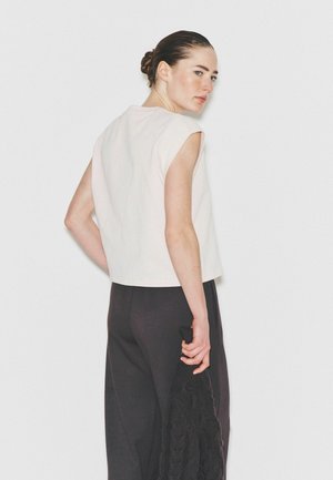 Kvinde med håret sat op i en knold iført hvid ærmeløs top og sorte bukser, der vender ryggen til og holder en sort strikket trøje i hånden.