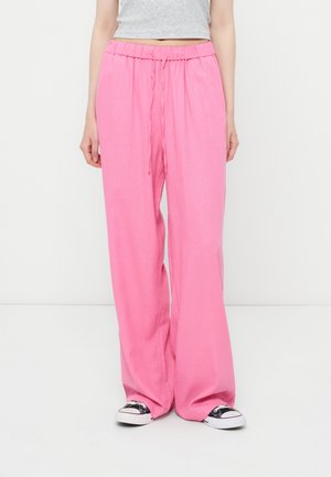 Γυναίκα που φοράει φαρδιά ροζ παντελόνια με κορδόνι και ασπρόμαυρα αθλητικά, στέκεται μπροστά σε λευκό φόντο.