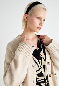 Beige neule cardigan, jossa on ribbestehmat, napit ja kaksi taskua, kerrostettuna mustan ja beige kuvioidun mekon päällä. Musta hiuspanta ja renkaat korvakoruina.