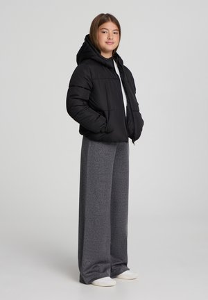 Giacca nera imbottita con cappuccio, zip frontale e pantaloni grigi larghi e texturizzati. Il modello è in piedi con le mani in tasca, indossando scarpe chiare.