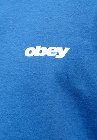 Μπλε μπλουζάκι από απαλό βαμβακερό ύφασμα με λευκή γραφική επιγραφή «obey» στο κέντρο του στήθους. Λείο ύφασμα χωρίς σχέδια.