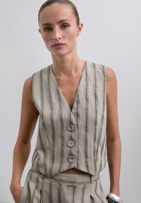 Kvinde iført en ærmeløs beige vest med lodrette striber og matchende højtaljede bukser, stående mod en ensfarvet lys baggrund.