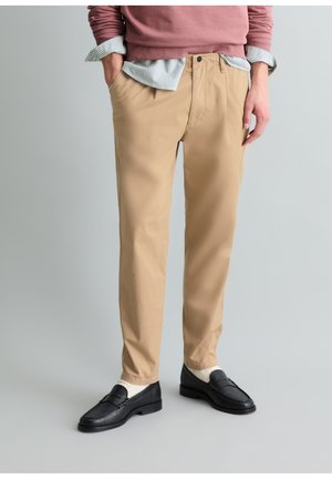 Mężczyzna w beżowych chino, czarnych mokasynach, białych skarpetkach, koszuli w paski i fioletowej bluzie, z rękami w kieszeniach na prostym tle.