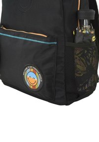 Mochila negra con un bolsillo con cremallera y un bolsillo lateral de malla. Cuenta con un parche circular con una cara sonriente y un diseño colorido.