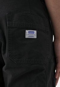 Pantalon en coton noir avec une poche à rabat ornée d'une étiquette blanche marquée "HUGO", affichant un texte bleu et une forme rectangulaire.