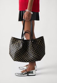 Svart og gull vevd totebag med to svarte håndtak, med en romslig kropp og en blank finish. Bæres med en rød genser og metalliske leiligheter.