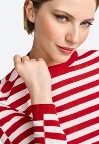 Rot-weißer gestreifter Pullover mit Rundhalsausschnitt, gerippten Bündchen und einer Nahaufnahme einer Hand, die auf dem Kinn ruht. Glatte Stofftextur.