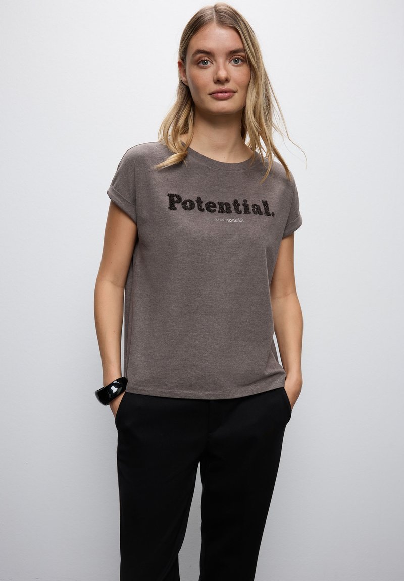 Brun bomullströja med korta ärmar, med texten "Potential." i svart på bröstet. Stylad med svarta byxor.