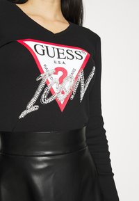 Musta ribattu pitkähihaisa paita, jossa on punainen ja valkoinen kolmio graafinen kuvasto, jossa lukee "GUESS" ja strassikoristelu, yhdistettynä kiiltävään mustaan nahkavyöhön.