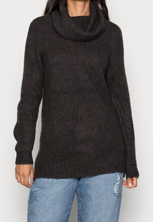 Struktureret sort strikket trøje med høj hals, lange ærmer og ribmønster, parret med slidte lyseblå jeans.