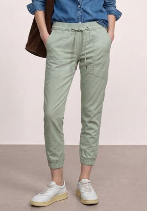 Personne portant un pantalon de jogging vert clair avec cordon, des baskets blanches, une chemise en denim bleu aux manches retroussées, et portant un sac marron à l'épaule.