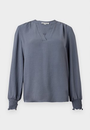 Långärmad, ljusblå blus med V-ringning, förankrad med samlade detaljer vid axlarna och smockade manschetter för textur och detalj.