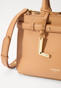 Tan leather handbag with a textured surface, gold-tone hardware, top handle, and detachable strap; features a gold "L" charm and logo.