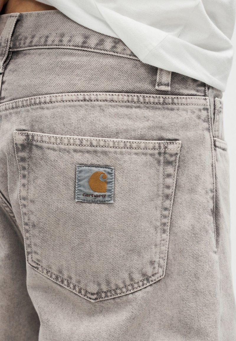 Vaqueros de denim gris claro con un bolsillo trasero con parche con el logo de Carhartt, usados con una camisa blanca metida dentro.
