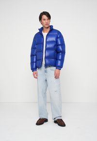 Zils puffer jaka ar horizontālu kviltu, augsts apkakle un rāvējslēdzējs; apvienota ar gaišām nolietotām džinsiem un brūnām kurpēm.